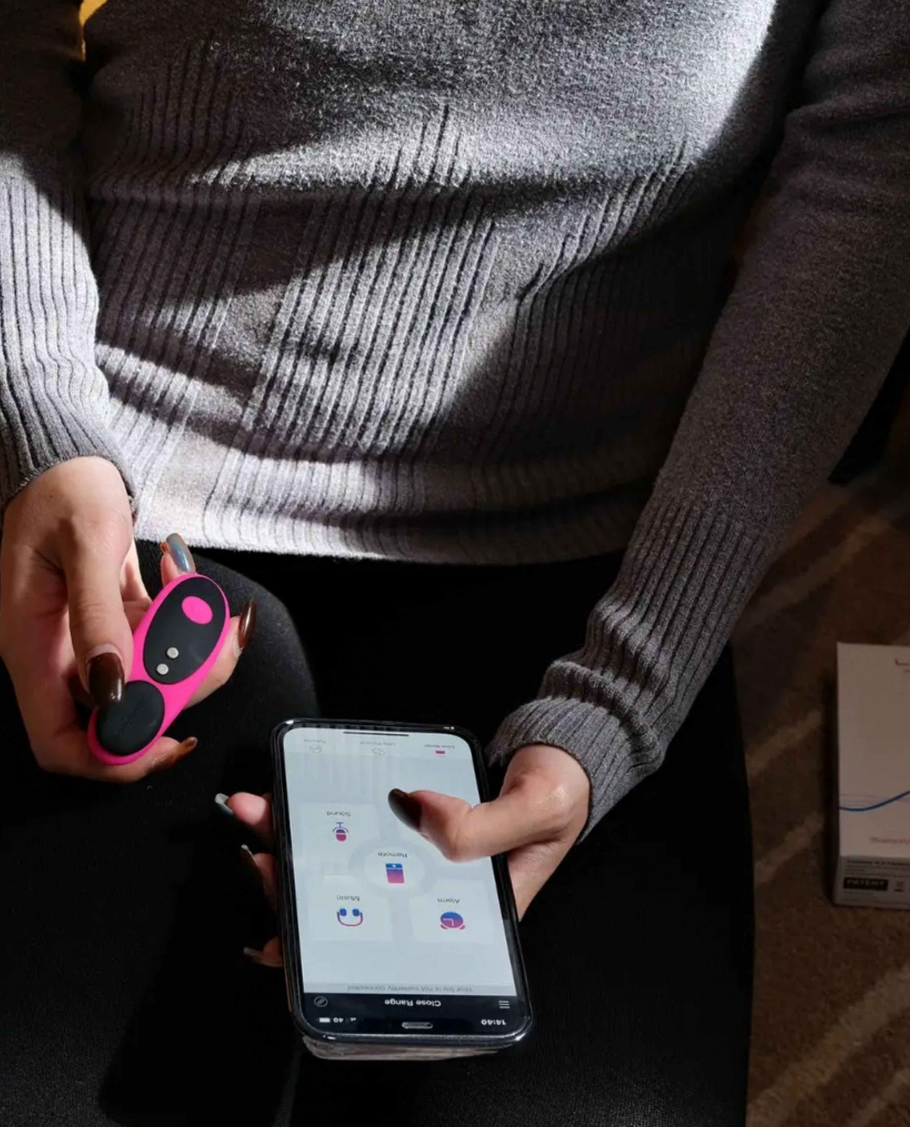 A person wearing a gray sweater holds a smartphone with the Ferri app-controlled panty vibrator, ready for some long-distance playtime.