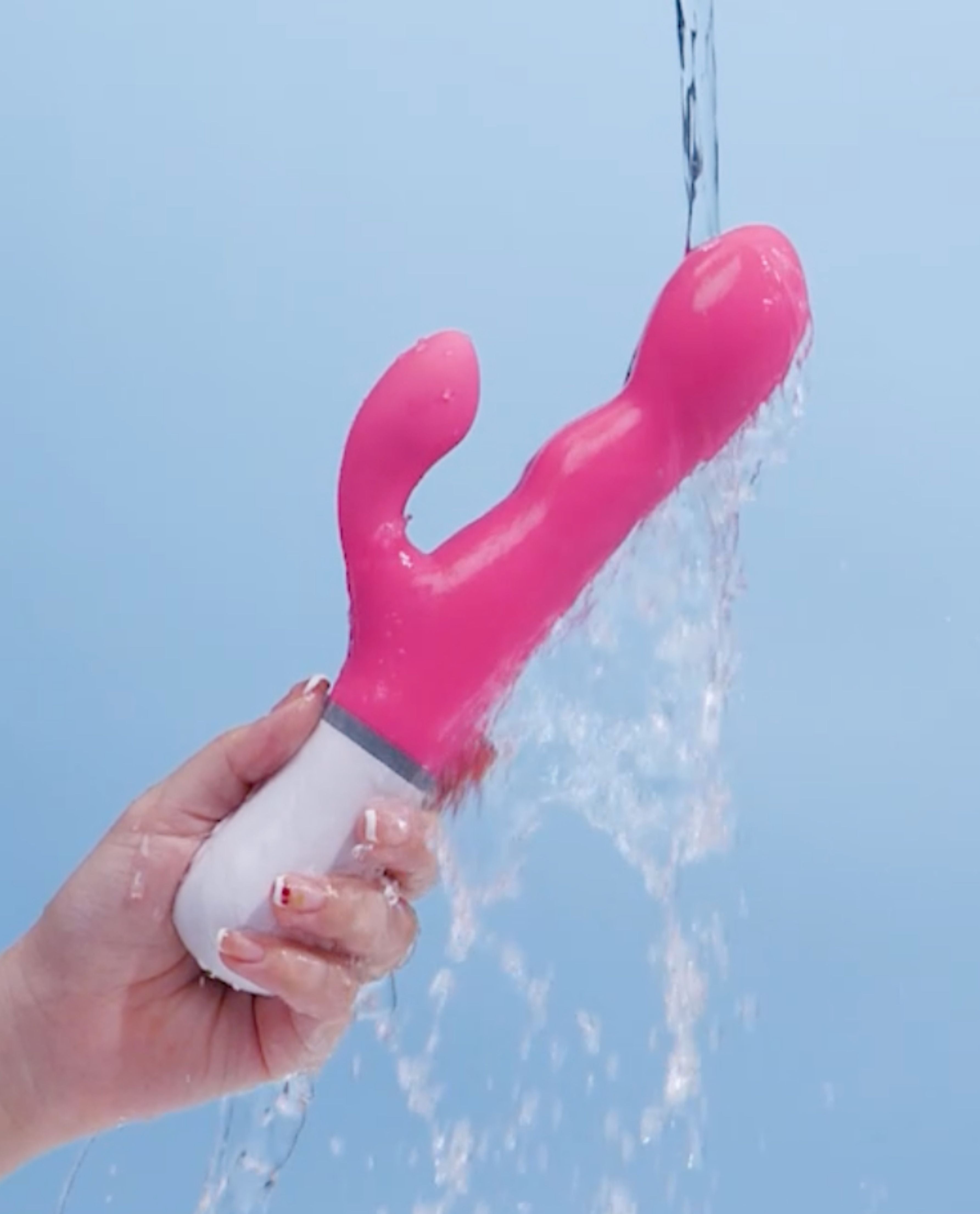 Nora by Lovense, a rotating rabbit dildo getting wet by a shower head held by a hand with water spraying from it against a clear blue sky.