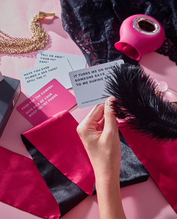 Box of Letz Get Naughty Cards, pink dare card, white truth card, pink silicone venus licking vibrator, gold chain tickler and satin blindfold on the table. Hand holding feather tickler