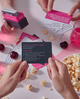 Box of Letz Get Naughty Cards, pink dare card, white truth card, popcorn and pink drink on the table