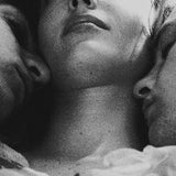 An intimate close-up black-and-white grain photo of a romantic throuple.A woman rests in the center with two faces gently pressed against her cheeks in a tender embrace, evoking themes of polyamory, love triangles, and aesthetic moodboards.