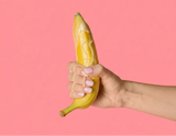 Hand holding a banana with white cream dripping over the peeled tip and down the fingers, against a pink background.