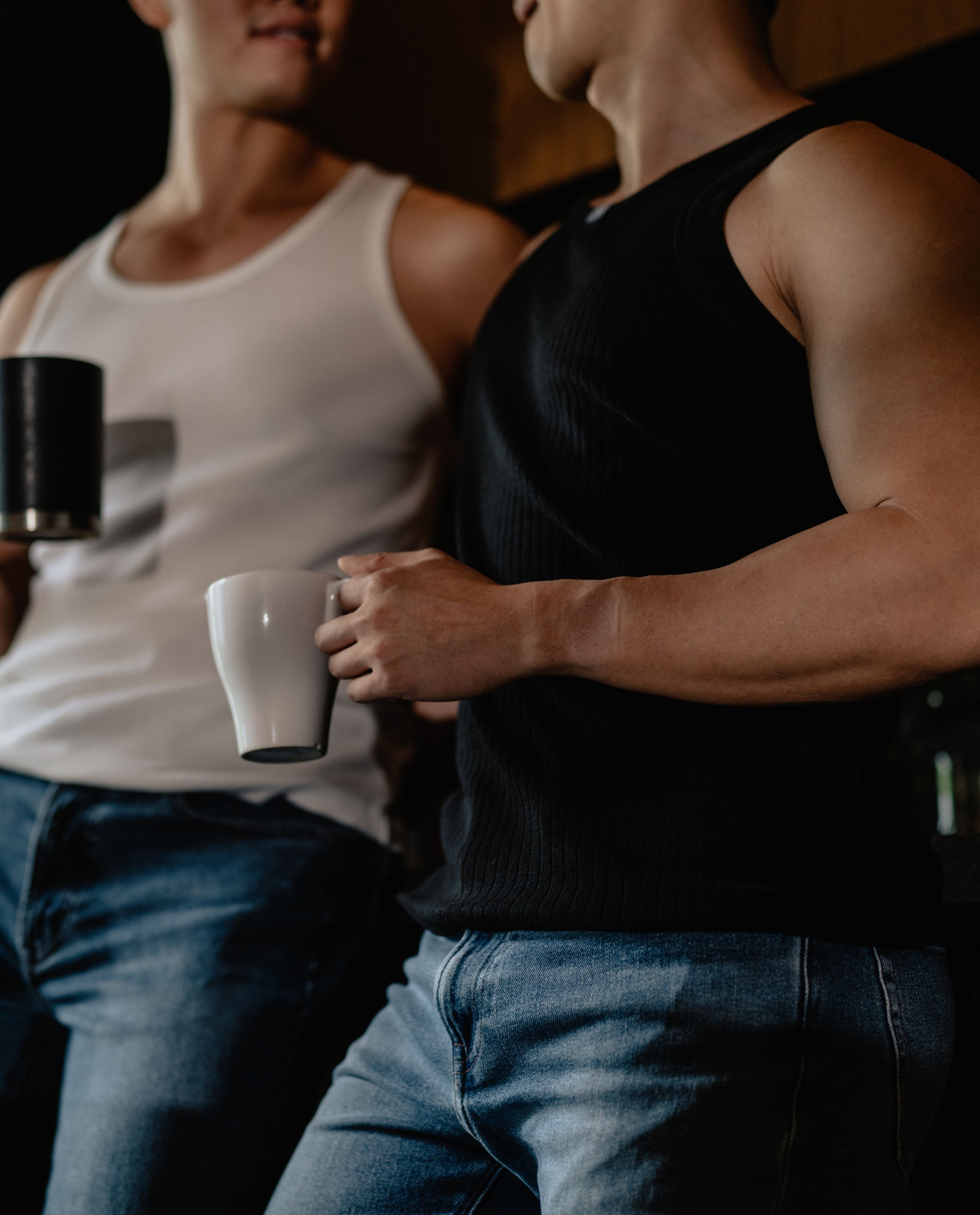 Two men holding coffee cups, one in a white tank top and the other in a black tank top.