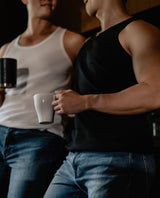 Two men holding coffee cups, one in a white tank top and the other in a black tank top.