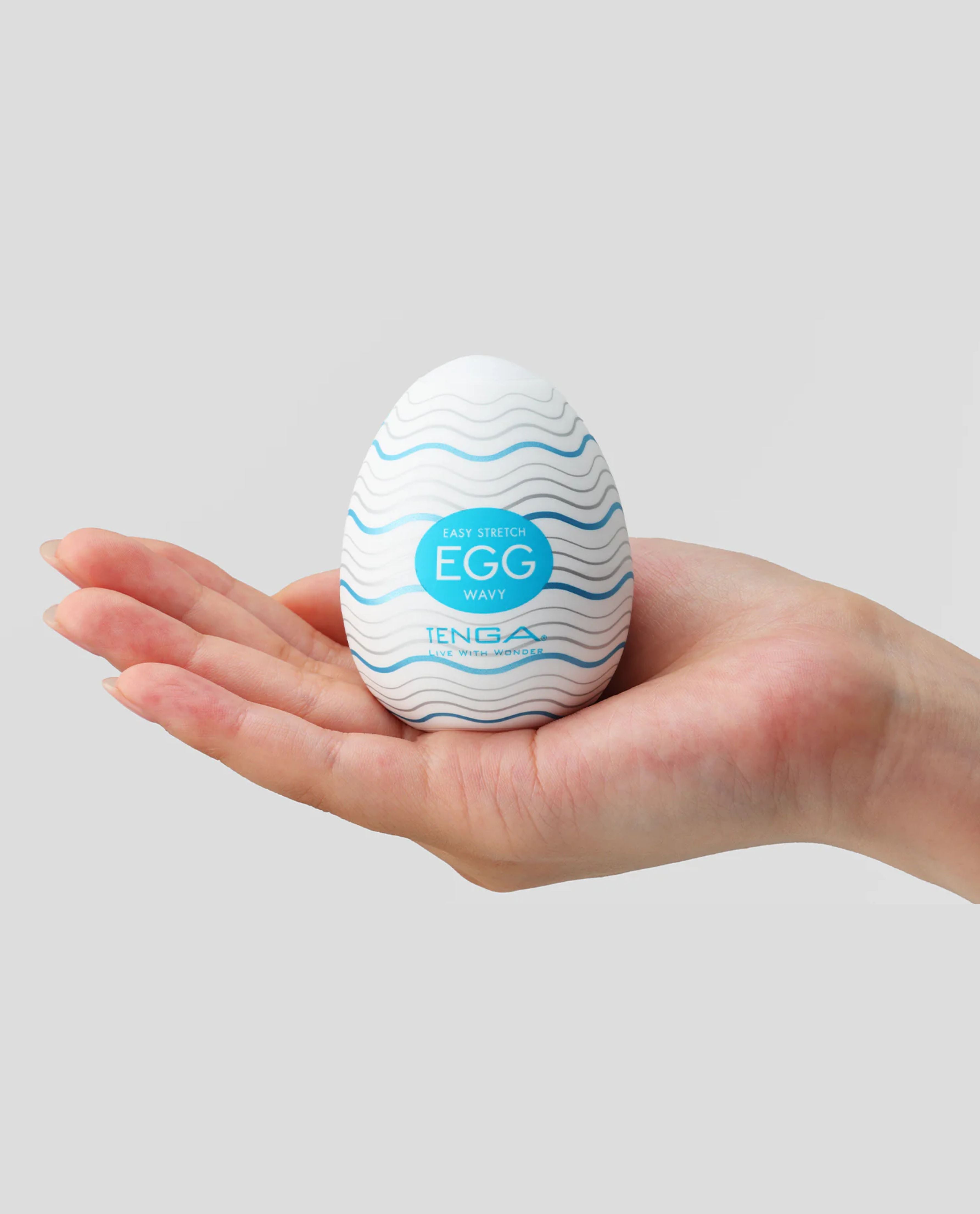 Hand holding a Tenga Wavy egg-shaped masturbator with blue text on a gray background