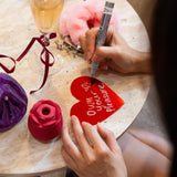 Person writing on a red heart-shaped card with a glass of champagne and flowers in the background.